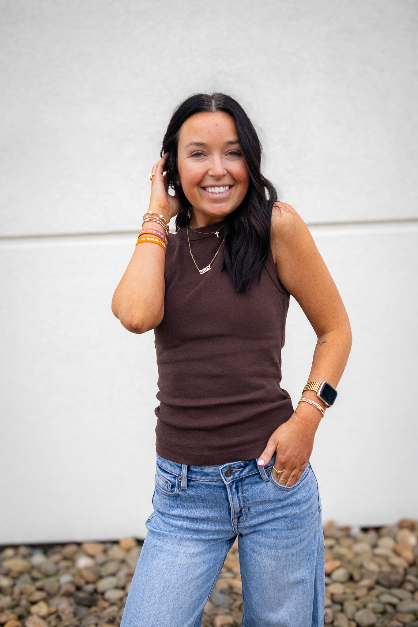 Woman wearing a brown sleeveless top and blue jeans against a white wall.