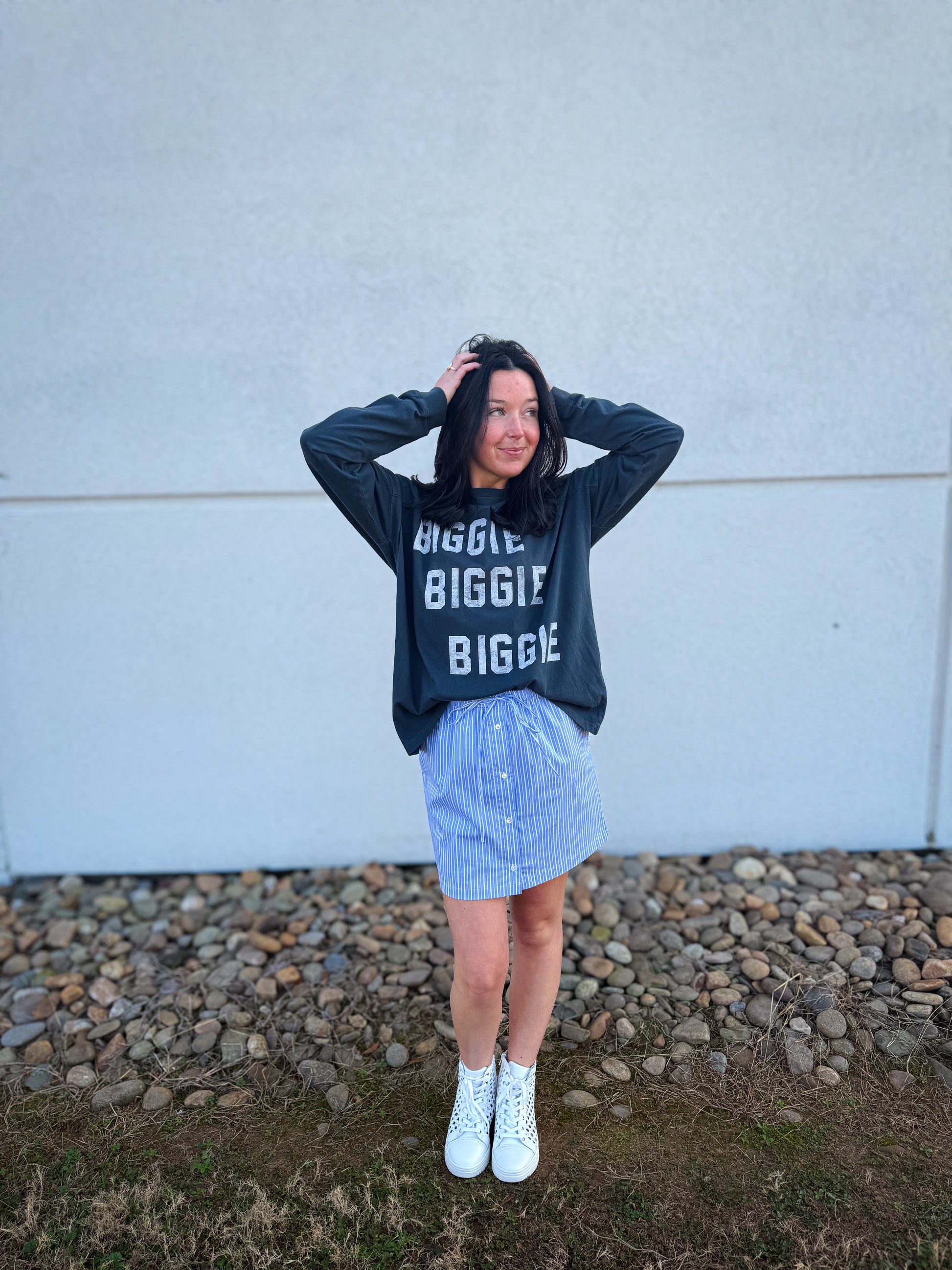 Person wearing a black sweatshirt with text, blue striped skirt, and white sneakers against a light-colored wall.