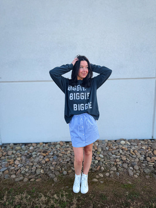 Person wearing a black sweatshirt with text, blue striped skirt, and white sneakers against a light-colored wall.