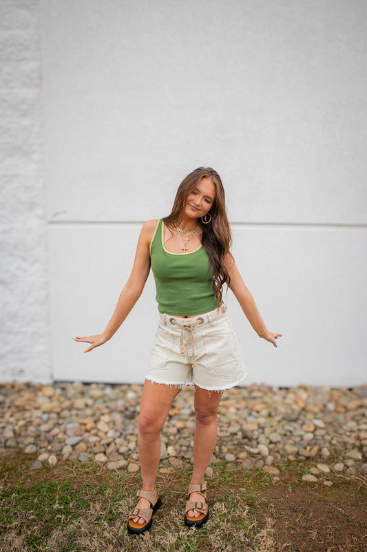 Woman wearing a green tank top and white shorts standing against a white wall.