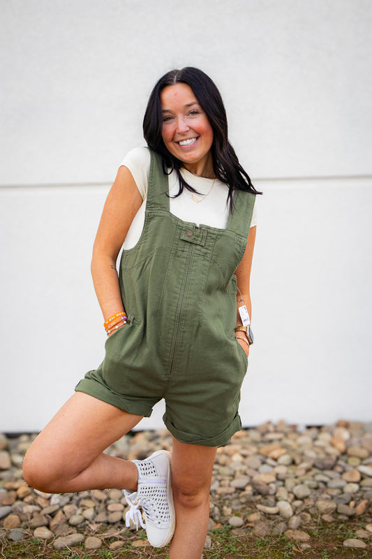 Woman wearing green overalls standing on a rocky ground with a white wall in the background