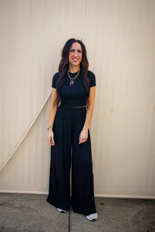 Woman wearing a black outfit with wide-leg pants against a beige wall.