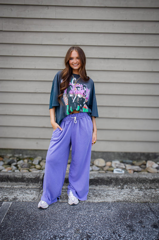 Woman wearing a graphic tee and purple pants standing against a gray wall.