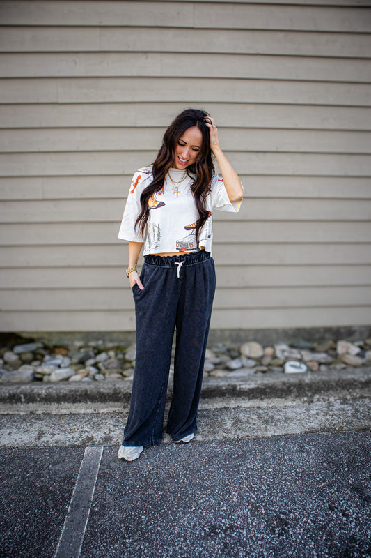 Woman standing on steps wearing a patterned top and black pants against a beige wall.