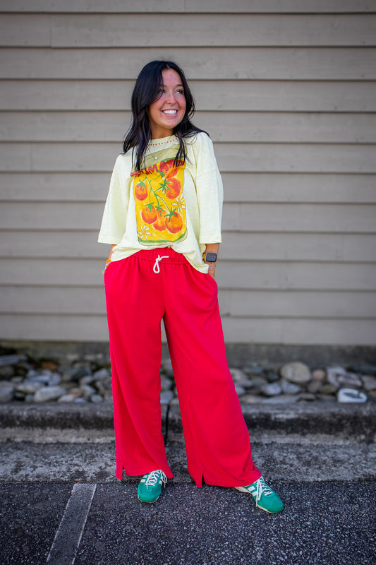 Person wearing a light-colored top with a colorful graphic and red pants standing against a gray wall.