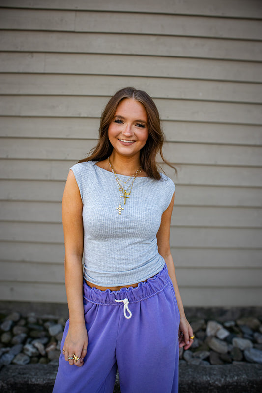 Woman wearing a light blue sleeveless top and purple pants standing against a beige wall.