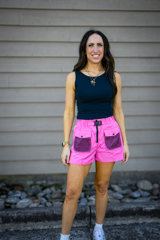 Woman wearing a black tank top and pink shorts standing against a neutral wall.