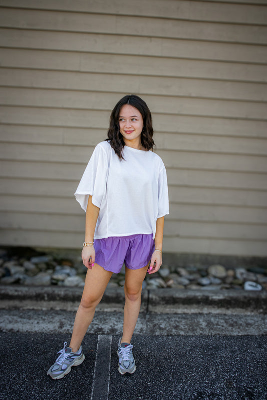 Woman wearing a white top and purple shorts standing against a beige wall.