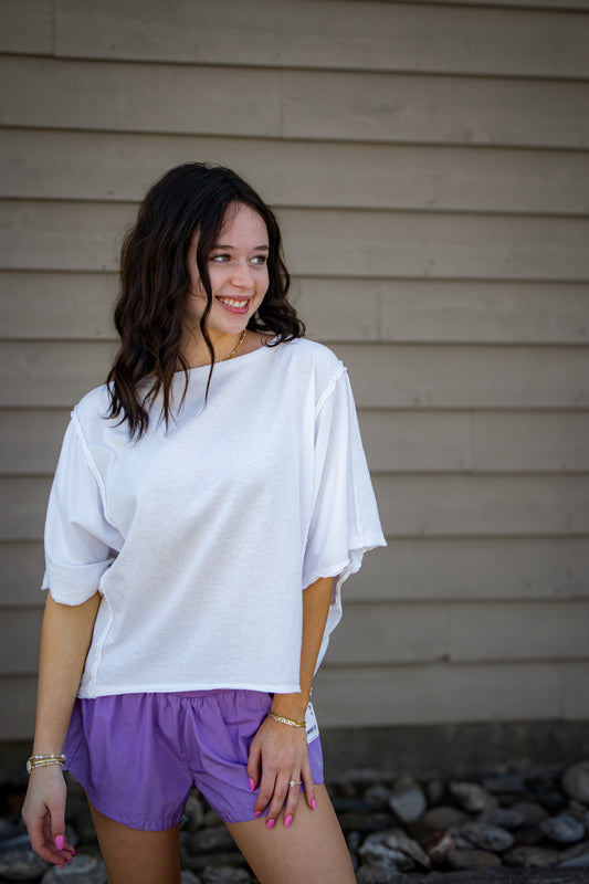 Woman wearing a white top and purple shorts standing against a beige wall.