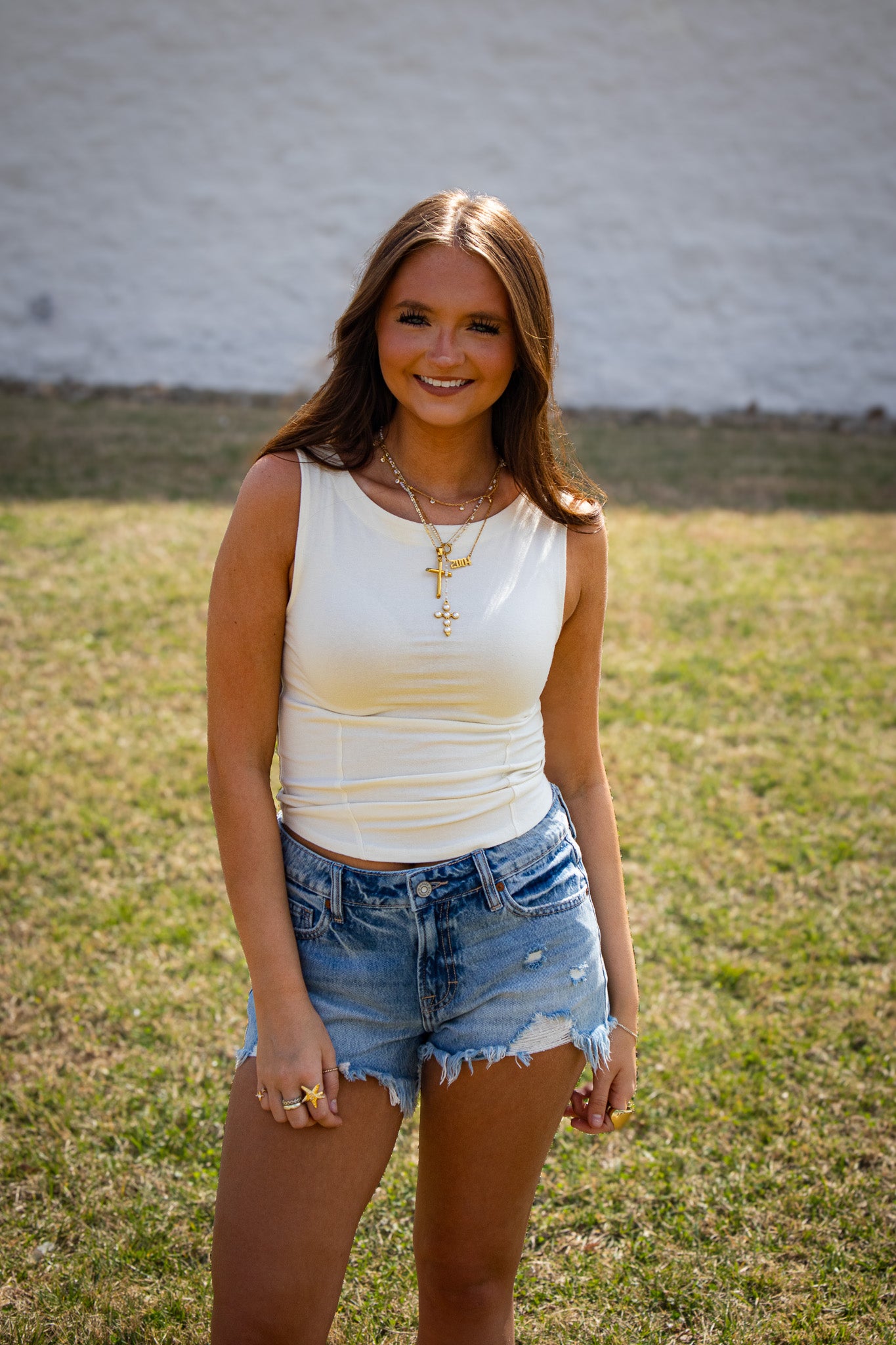 Woman wearing a white tank top and denim shorts standing in a grassy area with a white brick wall in the background.