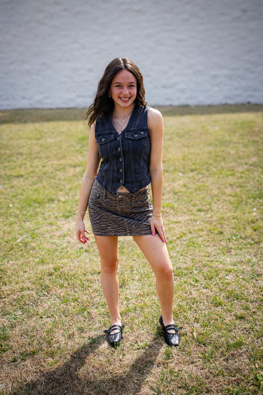 Woman standing on grass wearing a denim vest and patterned skirt.