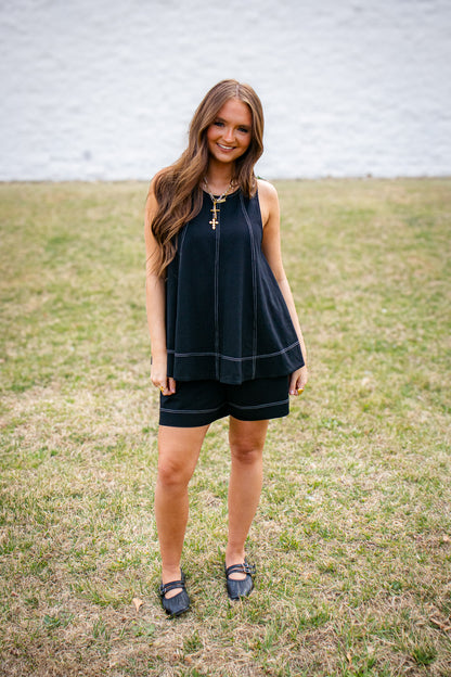 Woman wearing black sleeveless flowy top and matching shorts set with white stitching detail, styled with gold layered necklaces and black flats, casual chic outfit outdoors