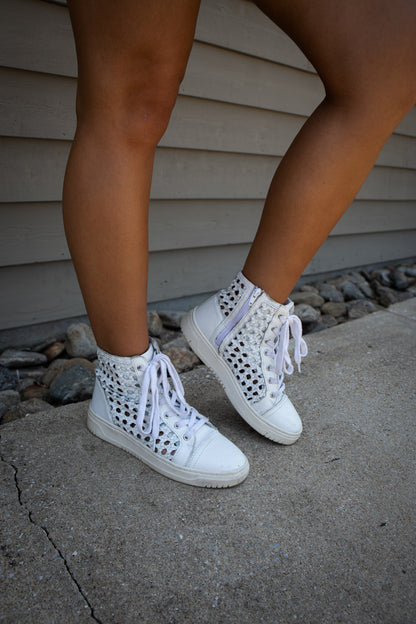 White high-top sneakers with perforated design worn by a person on a concrete surface.