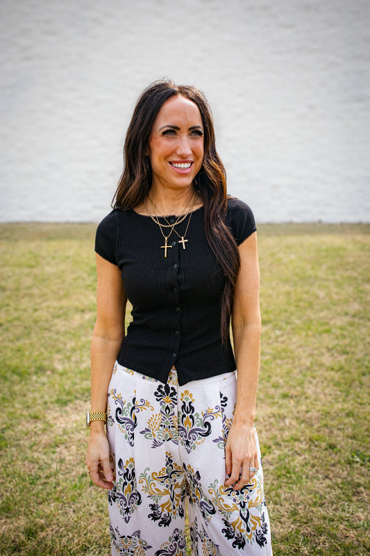 Woman wearing a black top and floral pants standing in a grassy area with a white wall background