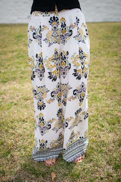 High-waisted wide leg pants with vintage-inspired floral damask print in cream, mustard, sage, and black, featuring a flowy fit and border detail hem, styled outdoors