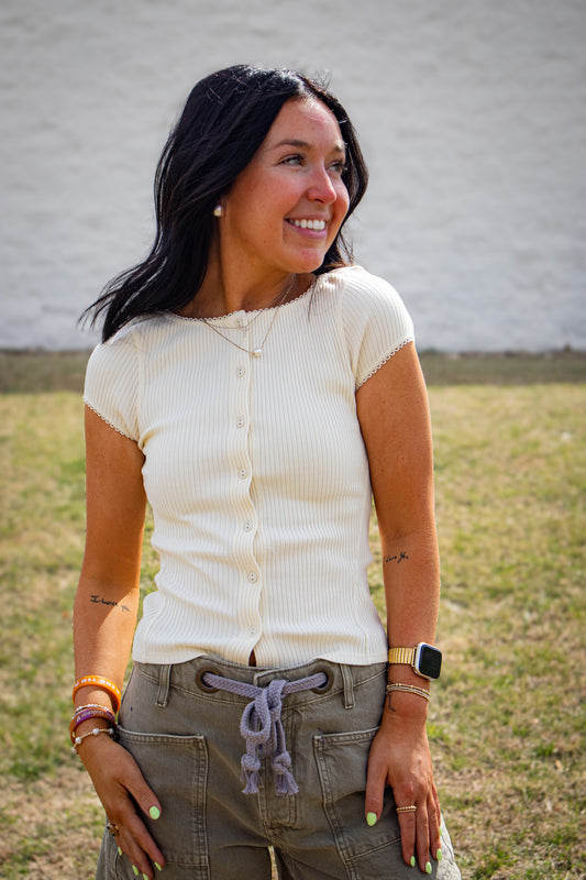 Woman wearing a cream-colored top and gray shorts standing outdoors against a white wall.