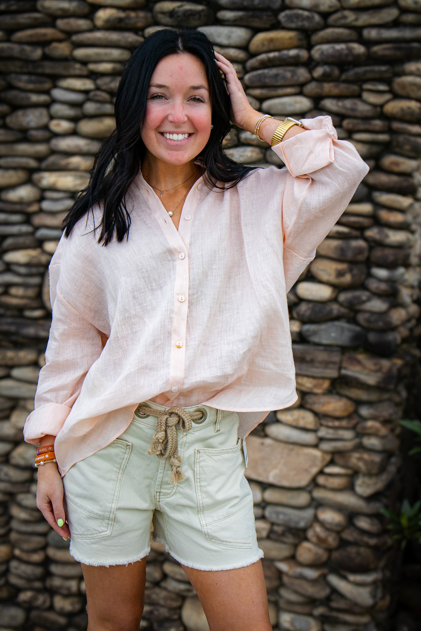 A smiling woman with dark hair stands in front of a stone wall, wearing a light pink button-up blouse and cream-colored drawstring shorts, with one hand touching her hair.
