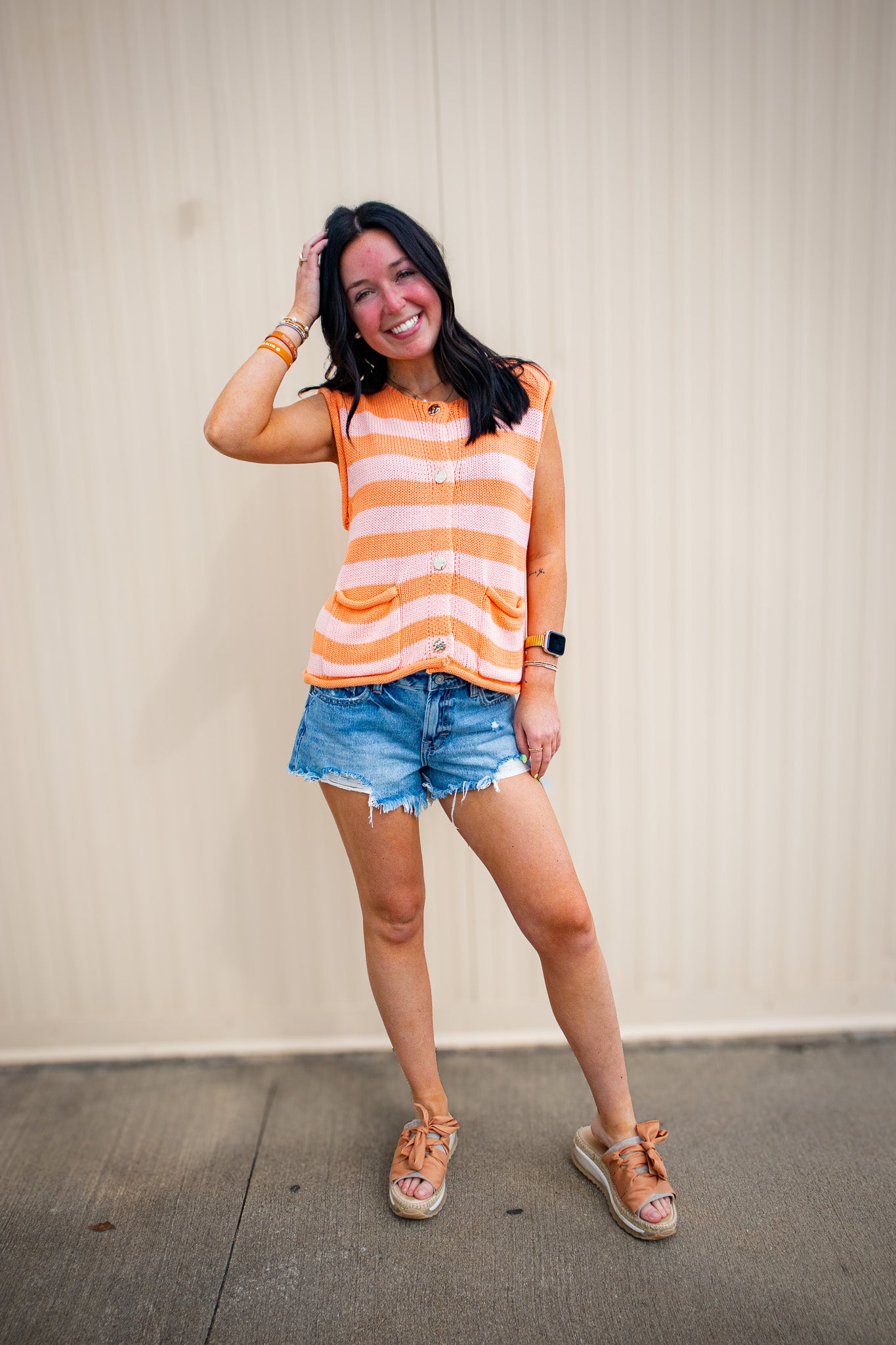 Woman wearing an orange and white striped sleeveless top with denim shorts against a beige wall.
