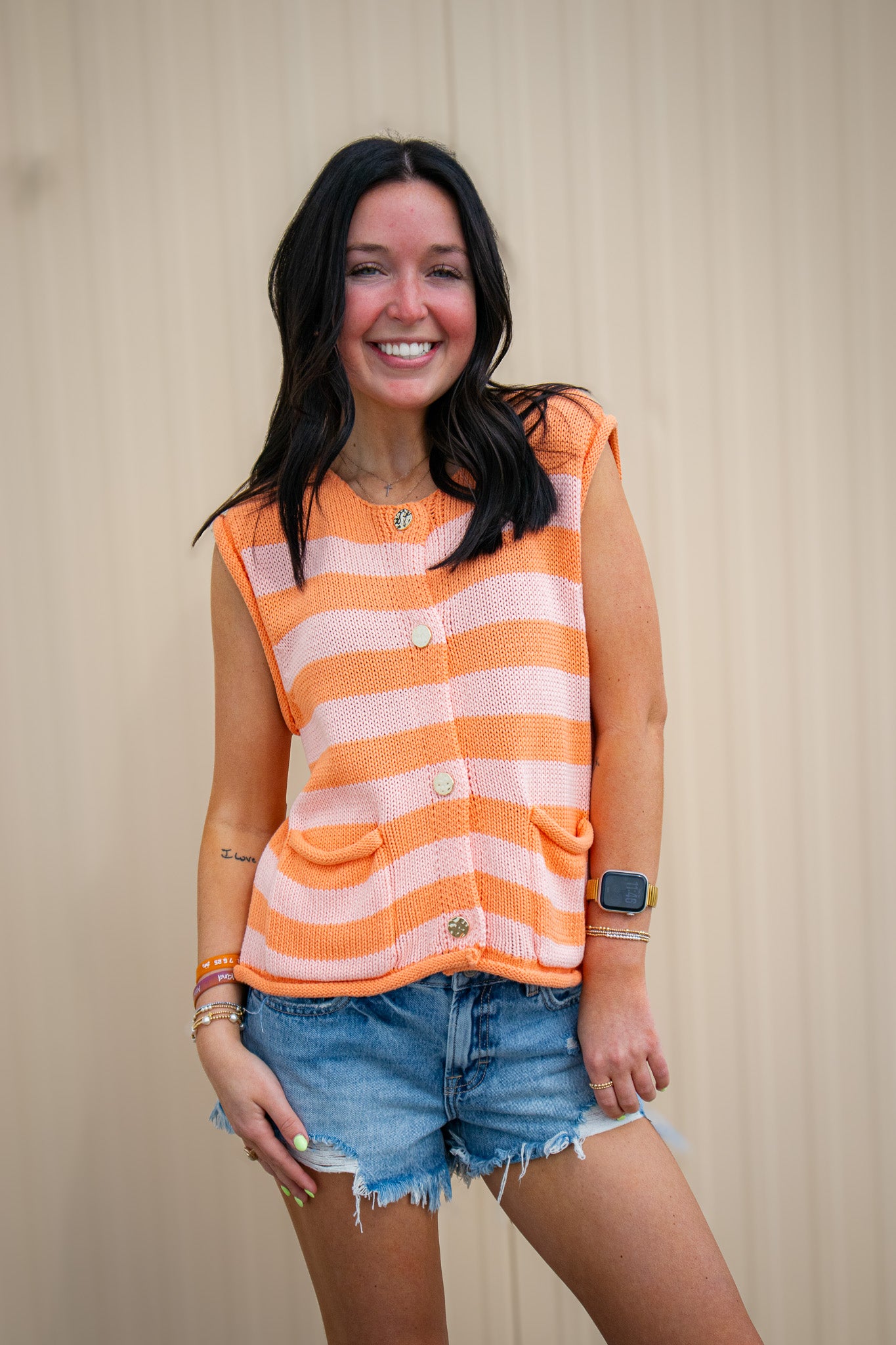 “A smiling woman stands against a neutral wall, wearing an orange and light pink striped sleeveless knit top with buttons and front pockets, paired with distressed denim shorts.