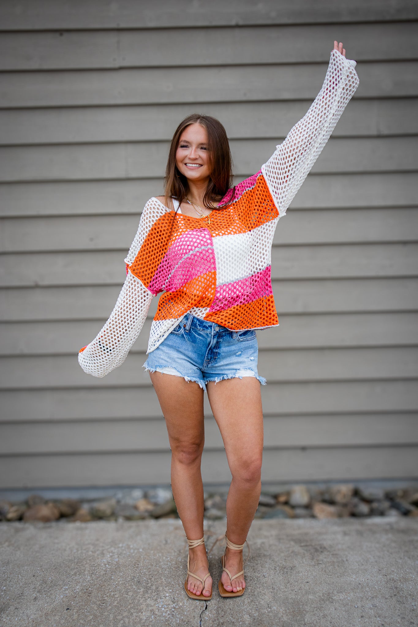 Woman wearing a colorful crochet sweater and denim shorts against a gray wall.