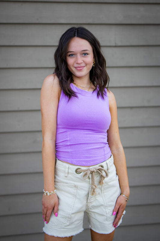 Woman wearing a purple top and beige shorts standing against a gray wall.