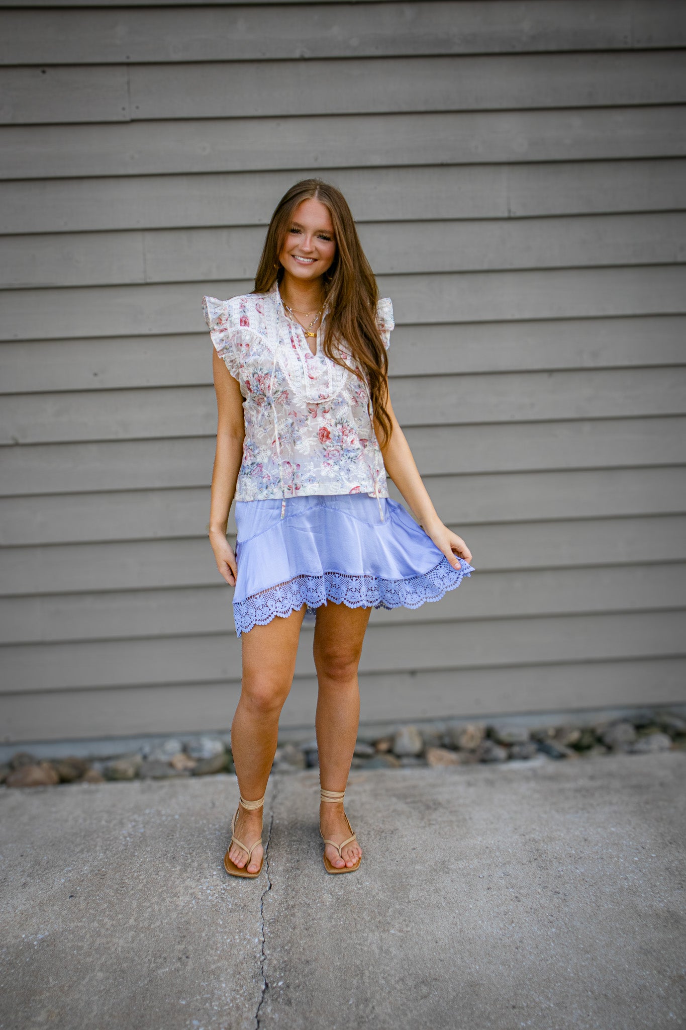 Woman wearing a floral top and purple skirt standing against a gray wall.