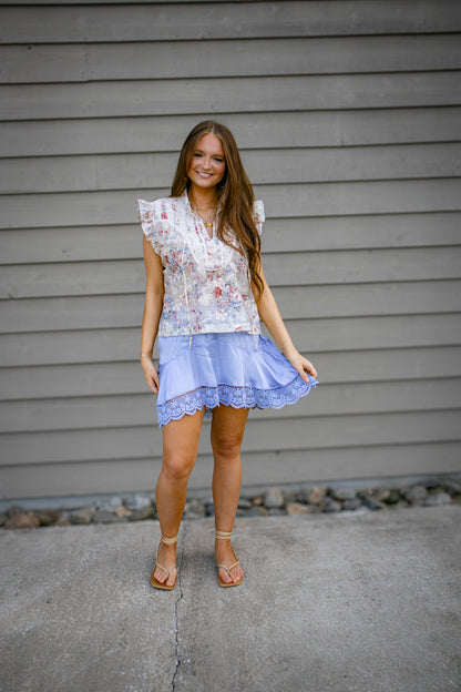 Woman wearing a floral top and purple skirt standing against a gray wall.