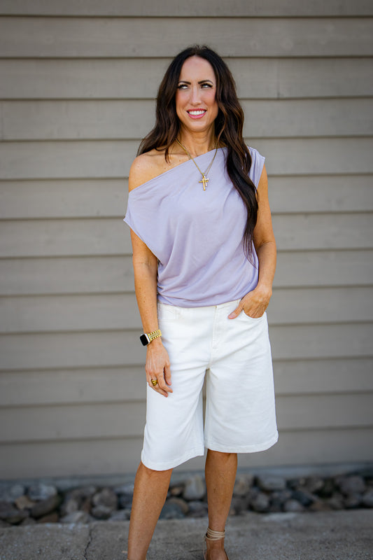 Woman wearing a light purple off-shoulder top and white shorts against a neutral background