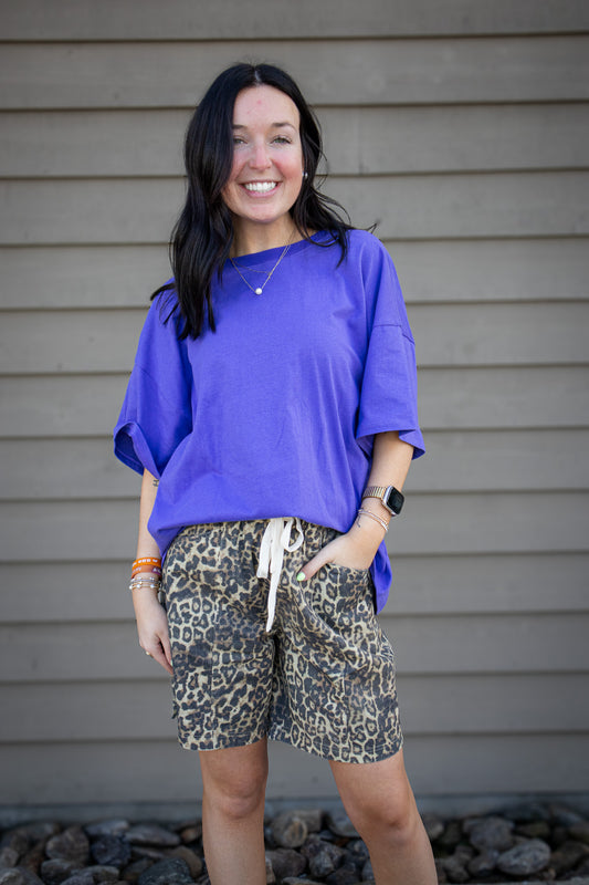 Woman wearing a purple shirt and leopard print shorts standing against a gray wall.
