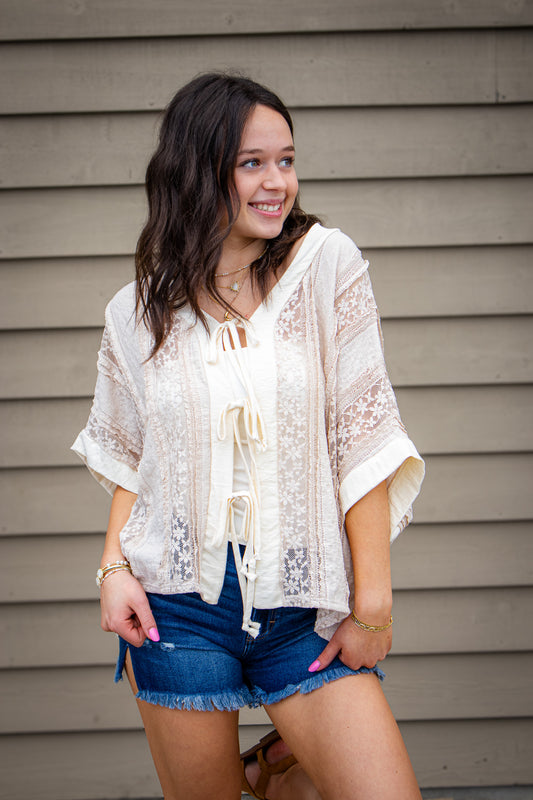 Woman wearing a tan lace top and denim shorts standing against a wooden wall.