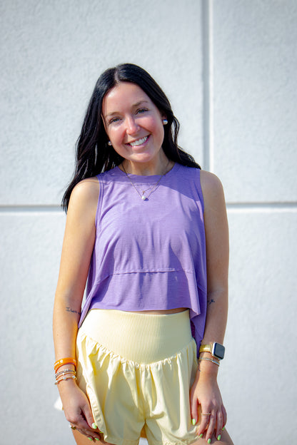 Woman wearing a purple sleeveless workout tank and yellow flowy shorts against a white wall