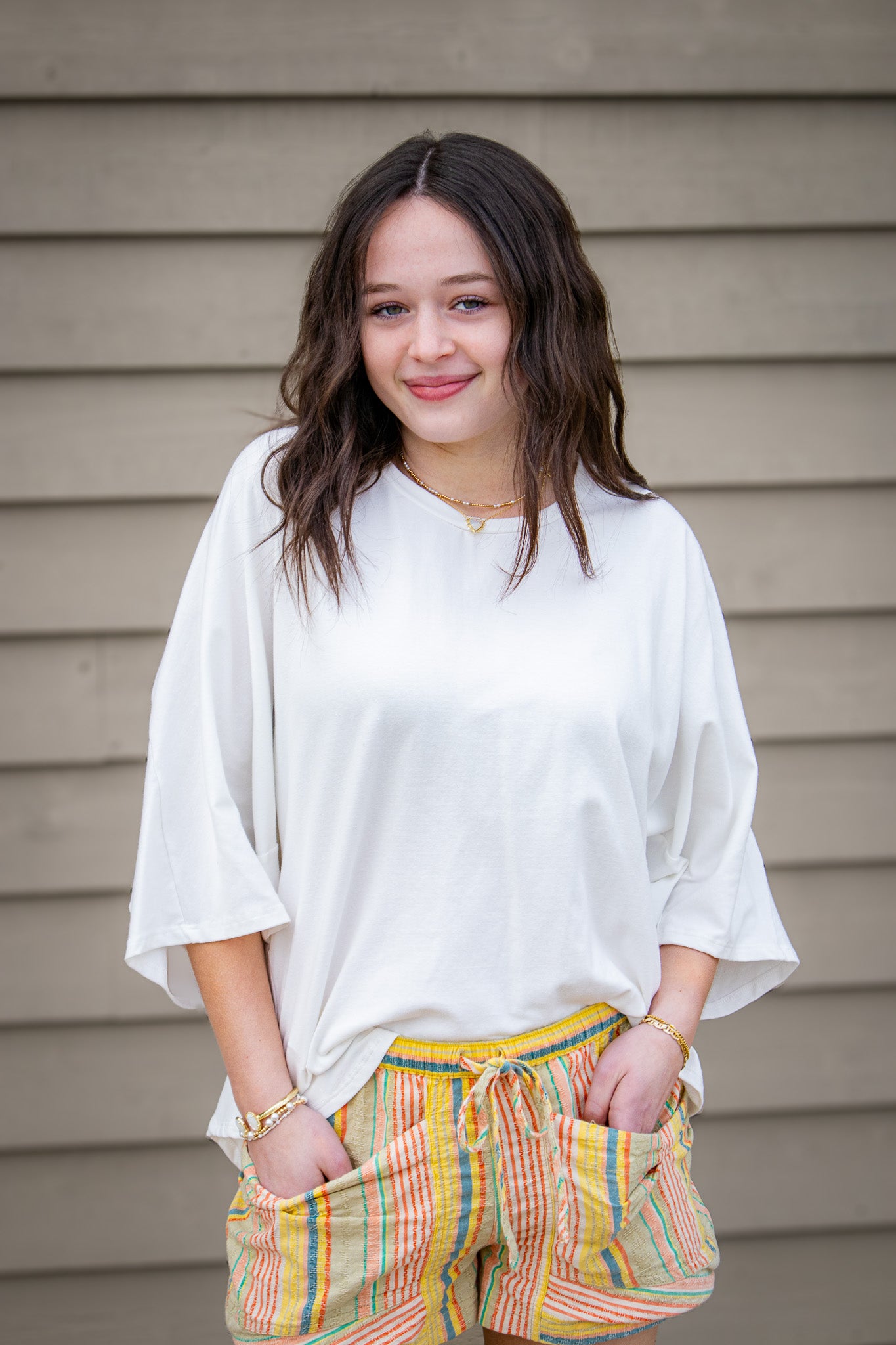 Woman wearing a white top and colorful shorts standing against a wooden wall.