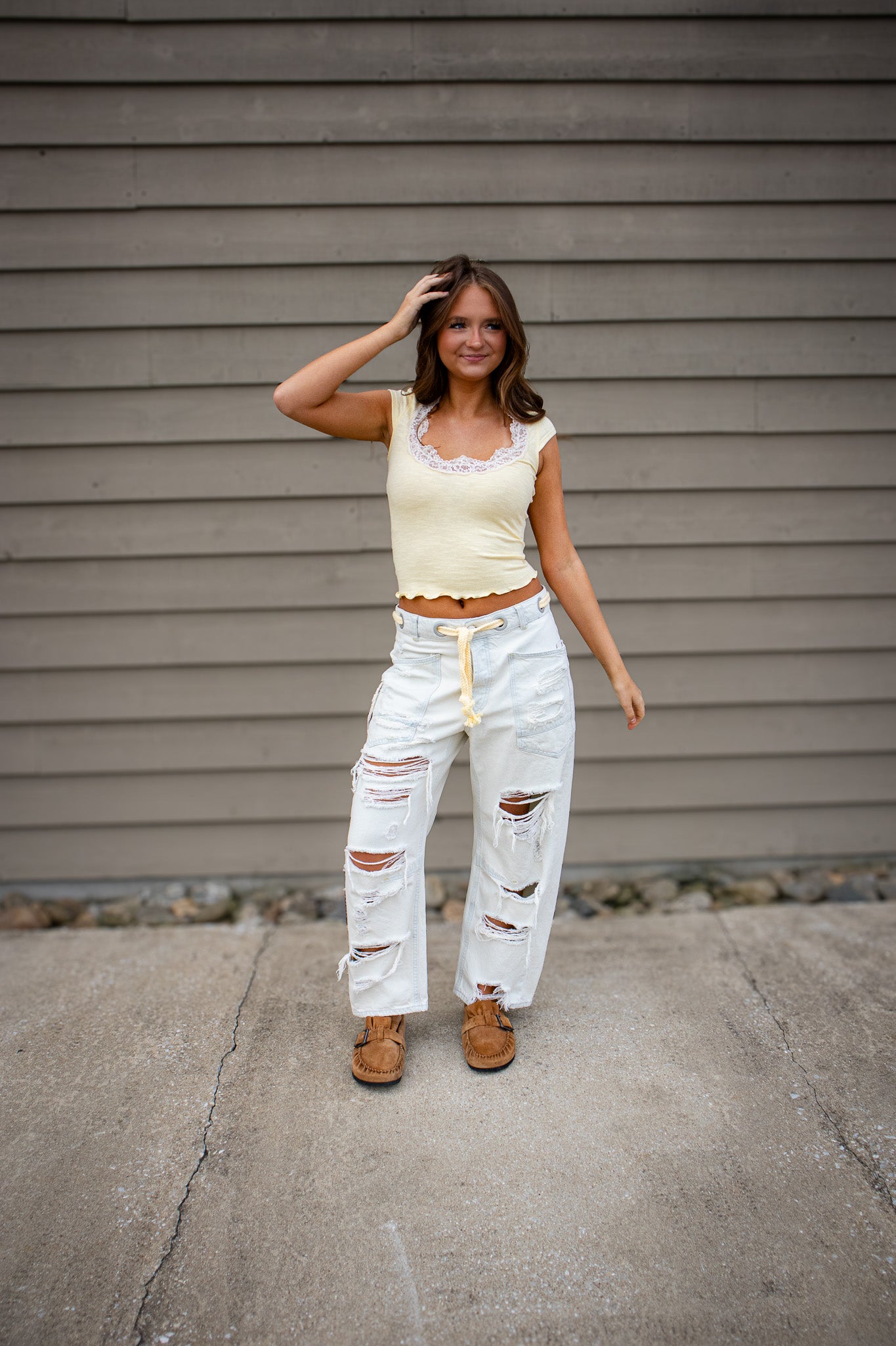 A young woman stands outdoors on a concrete surface in front of a gray siding wall, smiling slightly while touching her hair. She is wearing a light yellow cropped top with lace trim, distressed white wide-leg jeans with drawstring waist, and brown slip-on shoes