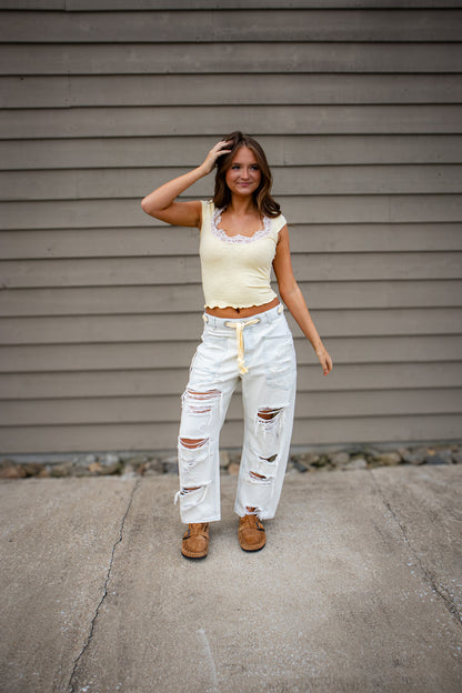 A young woman stands outdoors on a concrete surface in front of a gray siding wall, smiling slightly while touching her hair. She is wearing a light yellow cropped top with lace trim, distressed white wide-leg jeans with drawstring waist, and brown slip-on shoes