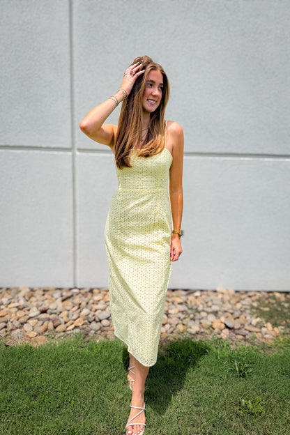 Model wearing a butter yellow eyelet midi dress with adjustable spaghetti straps, a flattering fitted bodice, and flowy skirt, styled with white strappy heels for summer weddings, brunch, vacations, and warm weather boutique outfits.
