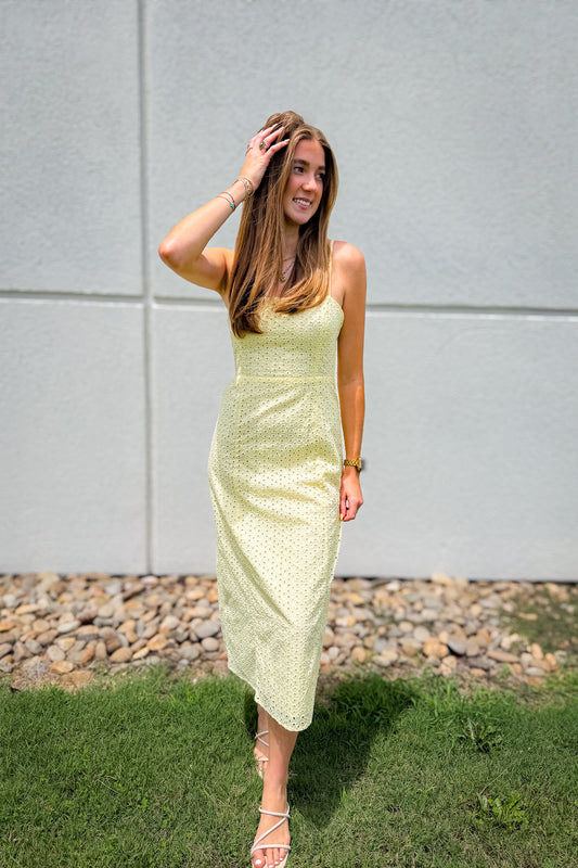 Model wearing a butter yellow eyelet midi dress with adjustable spaghetti straps, a flattering fitted bodice, and flowy skirt, styled with white strappy heels for summer weddings, brunch, vacations, and warm weather boutique outfits.