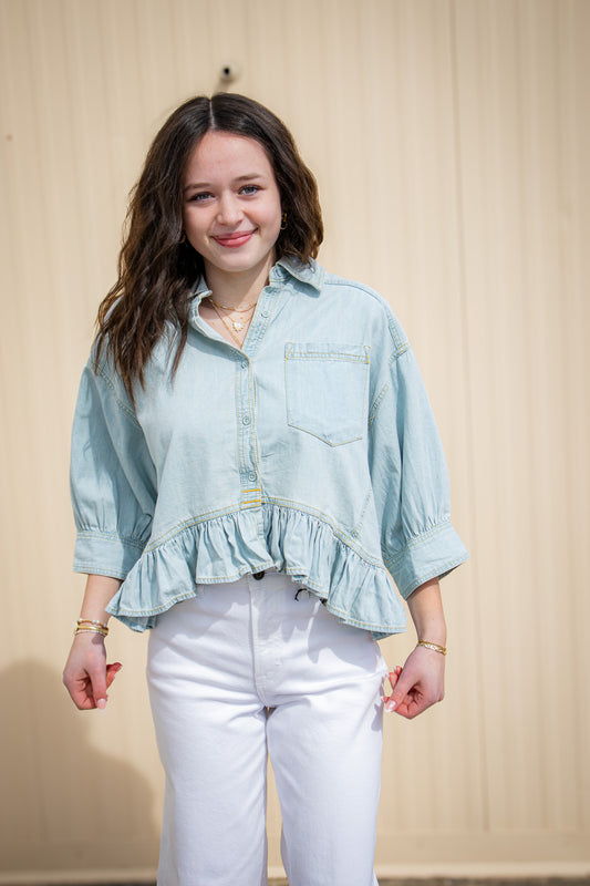 Woman wearing a light blue denim shirt with ruffled bottom half and white pants against a beige background