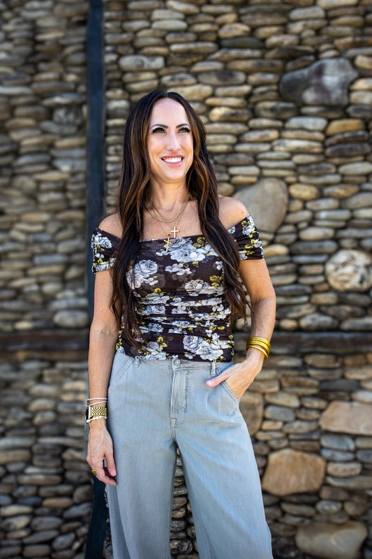 Woman wearing a floral off-shoulder top and light blue jeans standing against a stone wall.