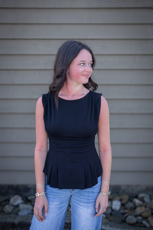 Woman wearing a black peplum top and blue jeans standing against a beige wall.