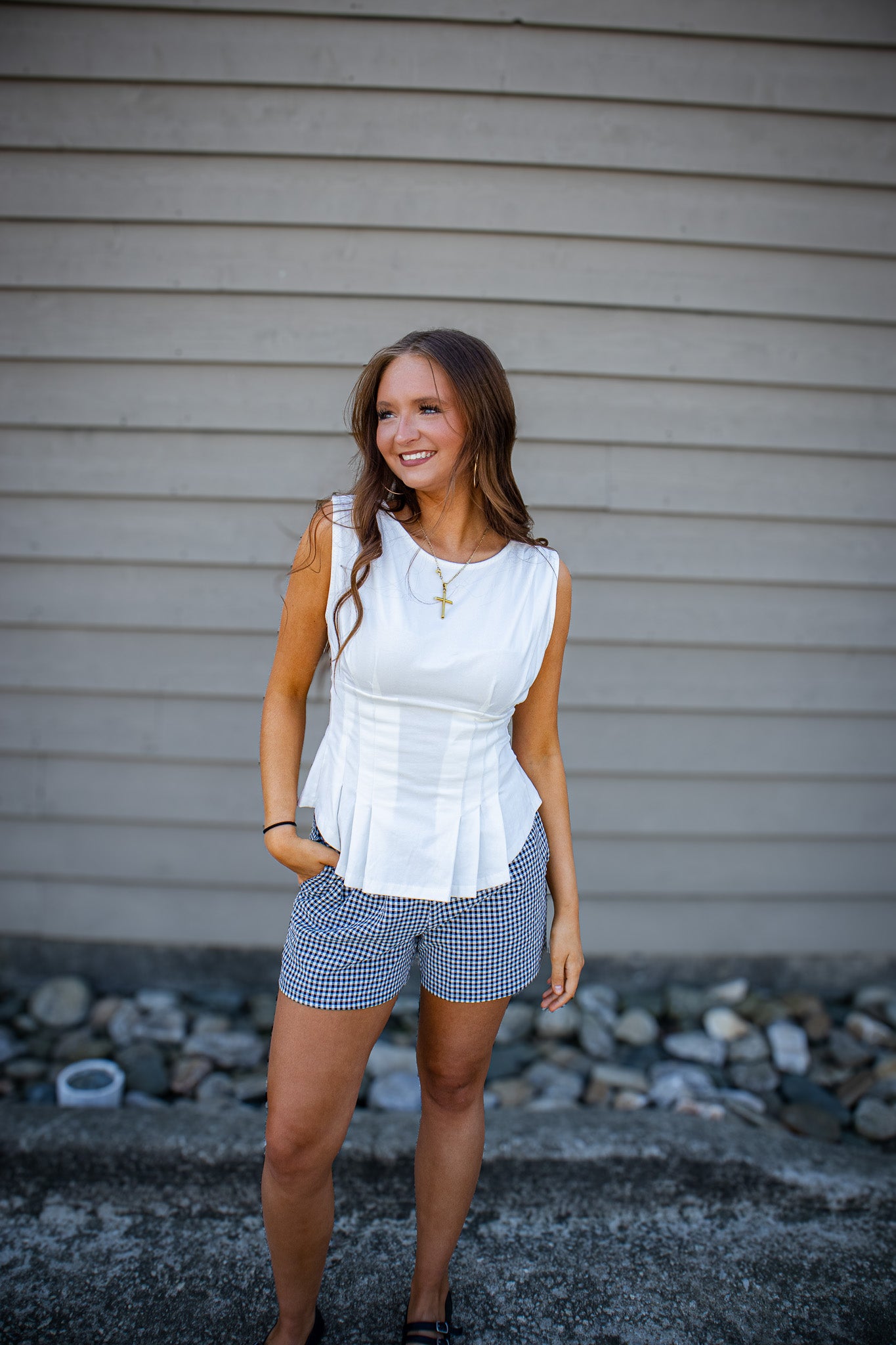 Woman wearing a white sleeveless top and checkered shorts standing against a gray wall.