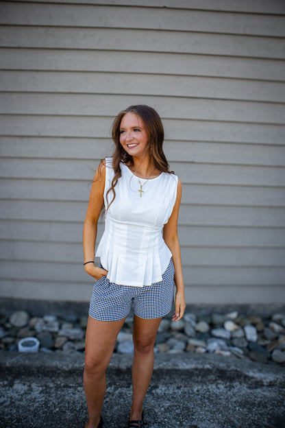 Woman wearing white sleeveless peplum top with black gingham shorts, casual chic summer outfit styled for trendy boutique fashion outdoors