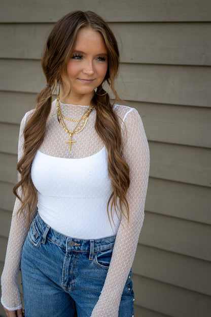 Woman wearing a white sheer top with a white tank underneath and blue jeans against a neutral background