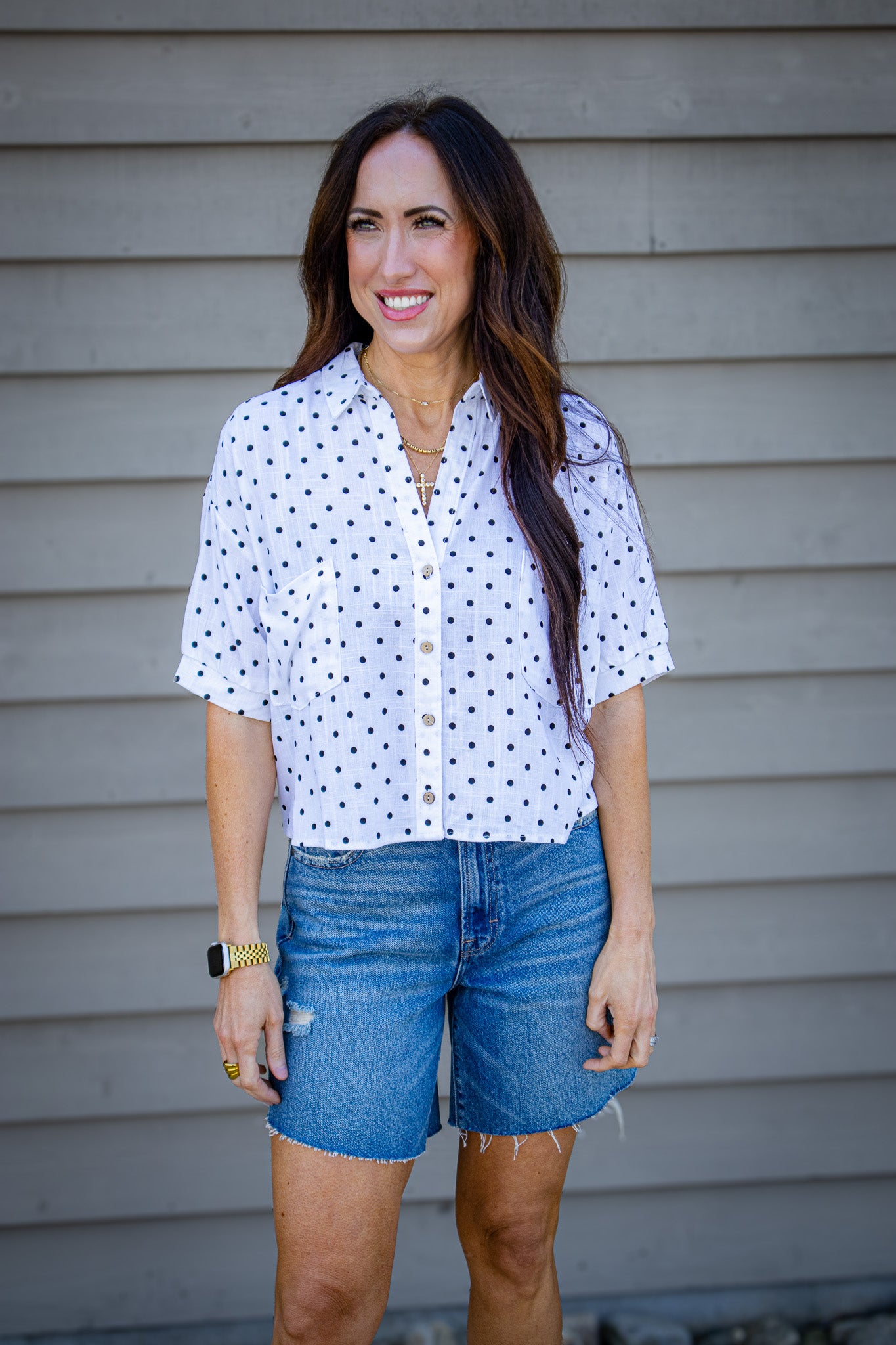 Woman wearing a white blouse with black polka dots and blue denim shorts standing against a gray wall.