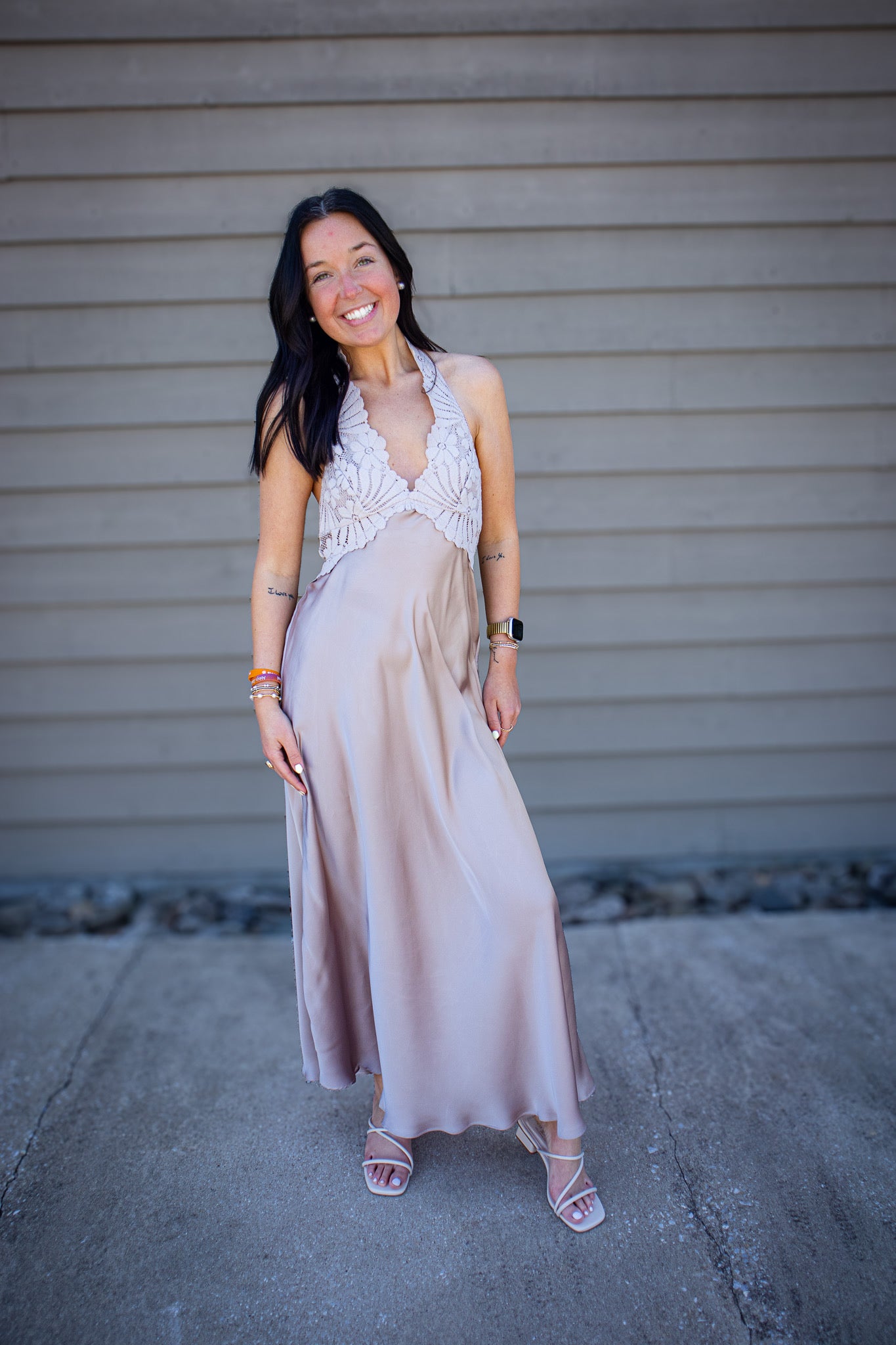 Woman modeling a neutral taupe maxi dress with lace halter neckline and flowy satin skirt, styled with strappy heels, casual outdoor fashion look