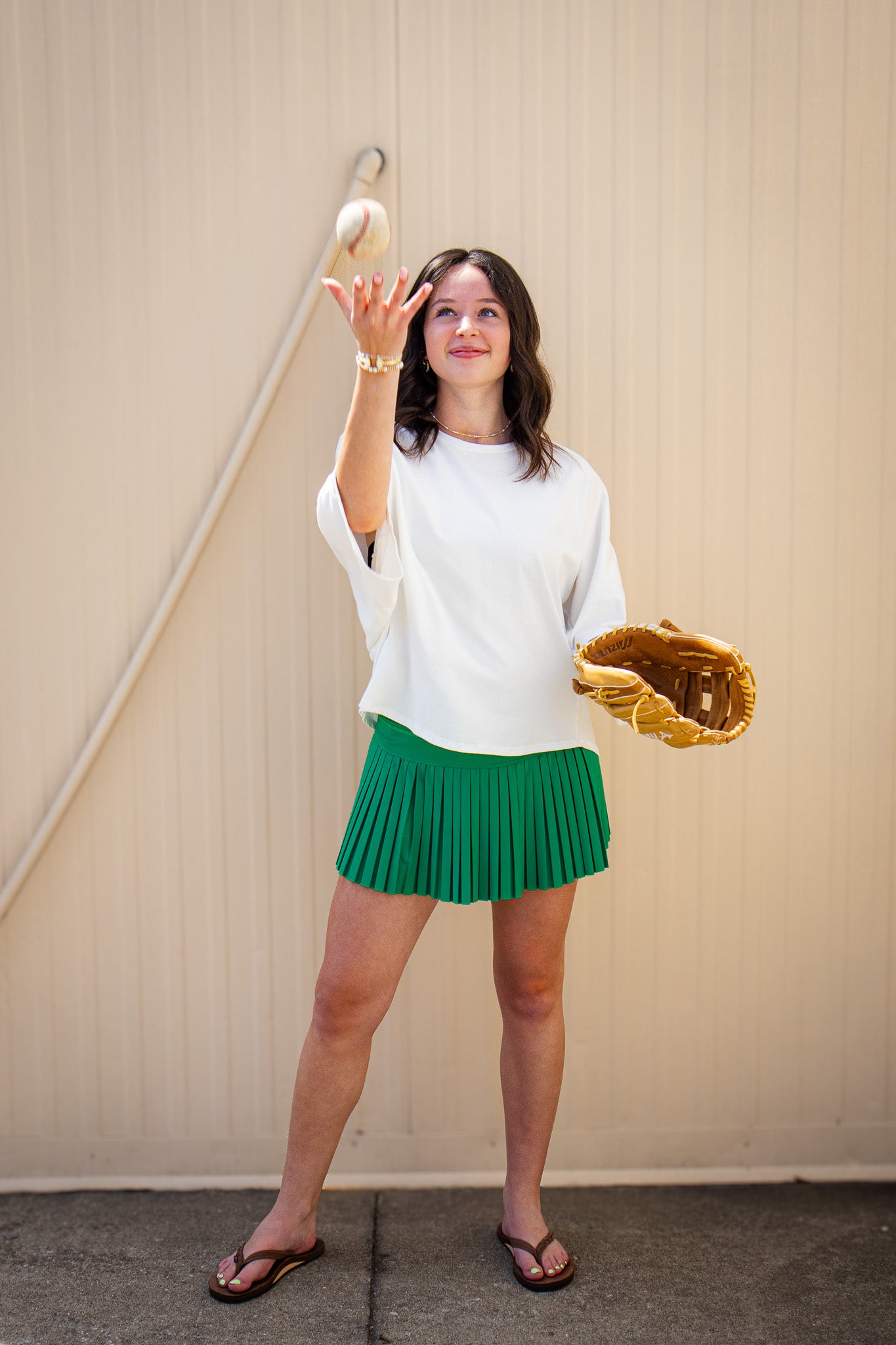 Model wearing a white oversized short sleeve top with a green pleated mini skort, brown flip flops, and gold jewelry, tossing a baseball while holding a leather glove for a sporty boutique game day outfit.