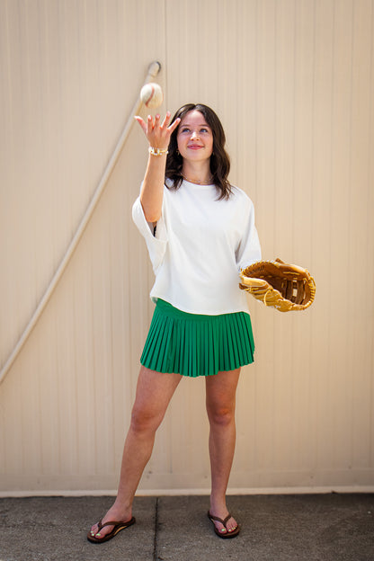 Model wearing a white oversized short sleeve top with a green pleated mini skort, brown flip flops, and gold jewelry, tossing a baseball while holding a leather glove for a sporty boutique game day outfit.