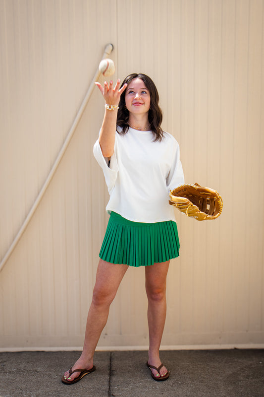 Model wearing a white oversized short sleeve top with a green pleated mini skort, brown flip flops, and gold jewelry, tossing a baseball while holding a leather glove for a sporty boutique game day outfit.