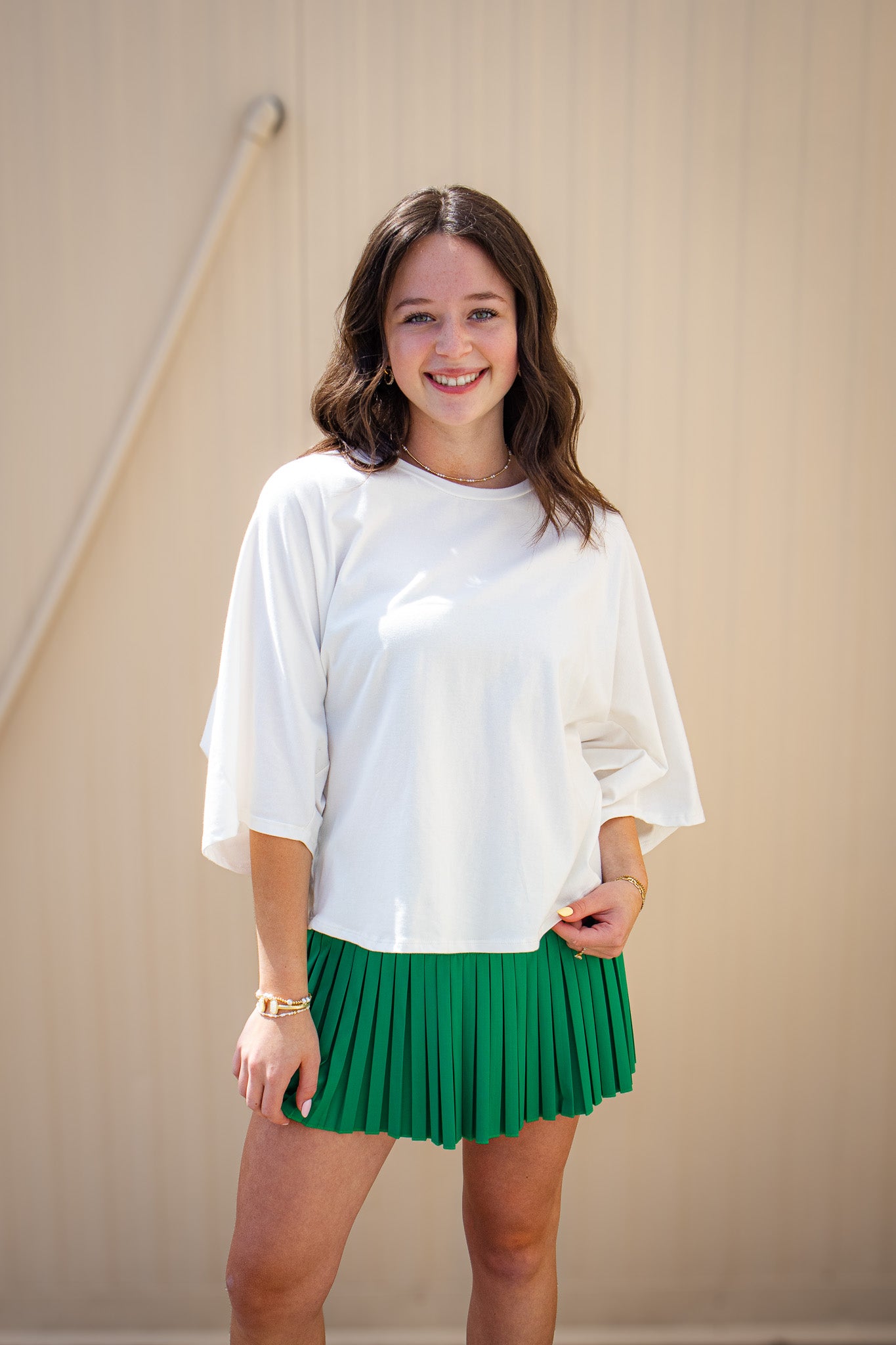 Model wearing a white flowy short sleeve top with an emerald green pleated mini skort, styled with gold jewelry for a cute sporty-chic boutique outfit perfect for game day, summer events, or casual weekends.