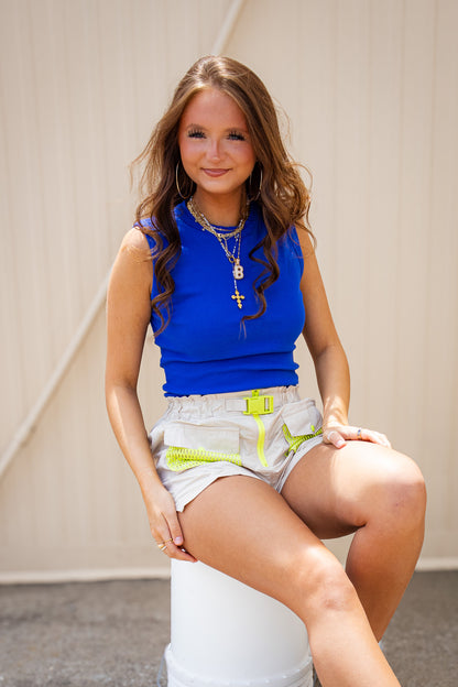 Model wearing a royal blue sleeveless fitted top with beige utility shorts featuring neon yellow mesh pockets and buckle detail for a bold women’s baseball game day outfit.
