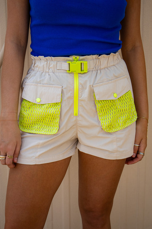Close-up of beige utility shorts with neon yellow mesh pockets, buckle belt, and zipper detail styled with a bright blue tank for a bold baseball game day outfit.
